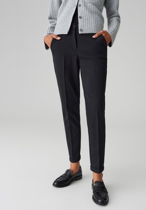 Schwarze maßgeschneiderte Hose mit schmaler Passform, gefertigt aus einem glatten Stoff. Verfügt über aufgerollte Säume, seitliche Taschen und wird mit schwarzen Mokassins kombiniert.