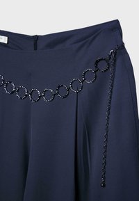 Pantalon bleu marine foncé avec chaîne décorative noire attachée, comportant de grandes maillons interconnectés et une chaîne prolongée suspendue sur le côté.