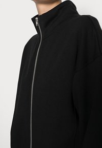 Sort zip-up jakke lavet af blødt stof, med høj krave og en sølvfarvet lynlås. Enkel og moderne design; ingen synlige mønstre eller dekorationer.