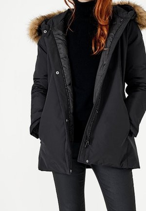 Nainen, joka kantaa mustaa talvitakkia, jossa on turkiskaulus, mustaa poolopaitaa ja mustia housuja, kädet takin taskuissa.