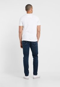 T-shirt blanc à manches courtes avec une étiquette carrée au dos, associé à un jean denim bleu foncé et des baskets blanches, vu de dos.