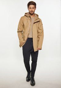 Beige vattentät jacka med huva, framdragkedja och två sidofickor, i kombination med mörka byxor och svarta snörstövlar. Slät textur.