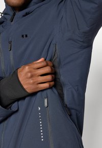 Giubbotto impermeabile blu scuro con tasca in rete con zip, accessori neri, dettaglio del logo e tessuto testurizzato per una maggiore comodità.