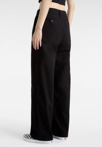 Svarta vida byxor i ett hållbart material, med en bakficka som stängs med knapp. Matchas med rutiga slip-on skor.