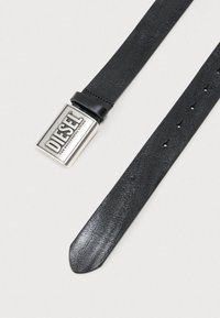 Cinturón de cuero negro con hebilla rectangular plateada grabada con el nombre de la marca. La correa tiene agujeros para ajustar la talla.