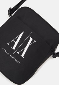 Czarna torba na ramię wykonana z tkaniny, z wyraźnym białym logo Armani Exchange, prostokątny kształt oraz regulowany pasek dla wygody.