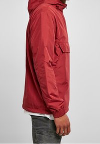 Osoba w czerwonej kurtce z kapturem, z elastycznymi mankietami, przednią kieszenią z klapką, białej koszuli pod spodem oraz ciemnych spodniach cargo, widok z boku.