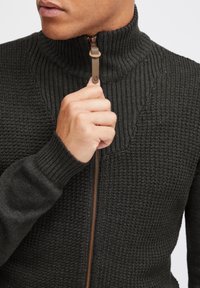 Suéter de punto acanalado de color gris oscuro con cuello alto y cremallera marrón. La tela parece tener textura, mostrando un ajuste ceñido y un diseño moderno.