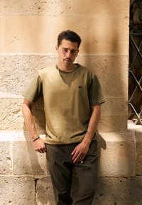 Maglietta verde oliva a maniche corte con un piccolo logo di elefante sul petto, abbinata a pantaloni verde scuro, in piedi contro un muro texturizzato.