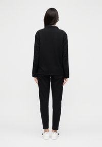 Zwarte sweatshirt en bijpassende broek van zachte stof. Heeft een losse pasvorm, lange mouwen en een minimalistisch ontwerp. Draagt witte schoenen.