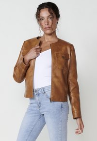Giacca di pelle marrone con scollatura rotonda, design aderente, chiusura frontale con zip e una singola tasca sul petto. Indossata sopra un top bianco.