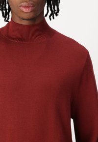 Burgundy színű kötött pulóver magas, bordás gallérral. Simaság jellemzi, hosszú ujjak, és laza szabású. Minimalista dizájn, látható minták nélkül.