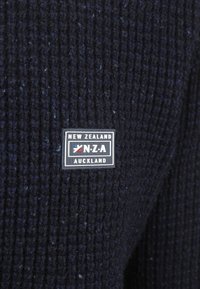 Donkerblauwe geribbelde gebreide stof met een klein rechthoekig label waarop staat "Nieuw-Zeeland N.Z.A Auckland" in witte en marineblauwe tekst.