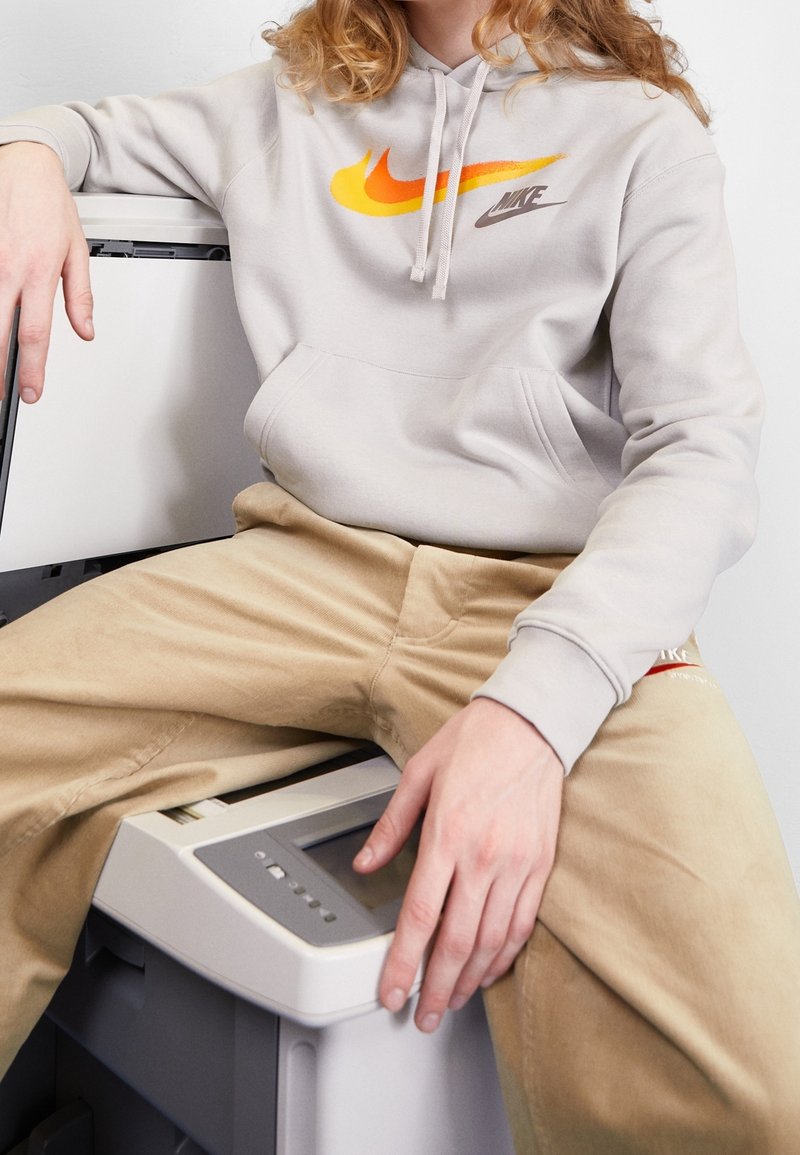 Ljusgrå huvtröja med orange och gul Nike-logotyp, ihop med beige byxor, sittande på en skrivare med en slät, matt yta.