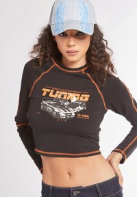 Maglietta nera a maniche lunghe e taglio corto con cuciture arancioni e stampa grafica di auto, abbinata a un cappellino grigio chiaro con dettagli di fiamme blu.