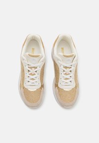 Beige og gull sneakers med en teksturert stoffoverdel, hvite snører og gummisåler, med et rundt logo på tungen.
