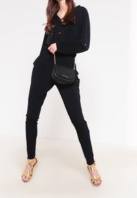 Černý overal s dlouhými rukávy a zapínáním na knoflíky vpředu, doplněný zlatými sandály na pásky a malou černou kabelkou přes rameno.