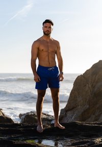 Blauwe zwembroek met een trekkoorddetail, staand op rotsen nabij de oceaan, heldere lucht op de achtergrond, zichtbare zeegolven.