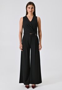 Tutina nera con top senza maniche di stile incrociato, pantaloni a gamba larga e una cintura con un dettaglio metallico circolare. Tessuto dalla texture liscia.