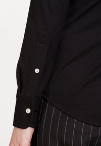 Black shirt with a textured fabric, featuring a buttoned cuff and a triangular cut at the sleeve. Paired with pinstriped pants.