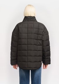 Geacă puffer neagră cu guler înalt, având pătrate quilted și mâneci largi. Are o croială lejeră și este confecționată dintr-un material neted.