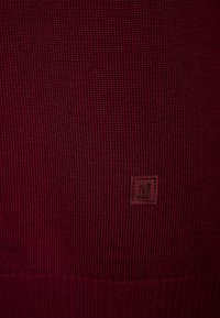 Burgunderfarbener Strickpulloverstoff mit gerippter Textur. Besitzt ein kleines rechteckiges Etikett mit der Buchstabe "M" in einer dunkleren Nuance.