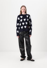 Maglione di lana blu con motivi a cuore bianchi, abbinato a pantaloni di pelle nera. La persona indossa una piccola borsa nera. Scarpe nere piatte completano il look.