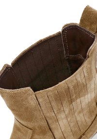 Close-up of a tan suede boot's elastic side panel with visible stitching and pull tab on the back.