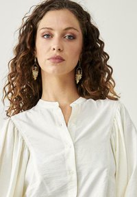 Camisa blanca con botón, de mangas abullonadas y cuello. La tela parece suave y ligera, con una textura sutil y un drapeado suave.