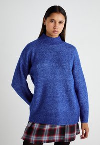 Modra volnena pulover s turtleneck ovratnikom in ohlapnim krojem, z rebrastimi manšetami in robom. Uporabljen z karirasto krilo v rdeči, črni in beli barvi.