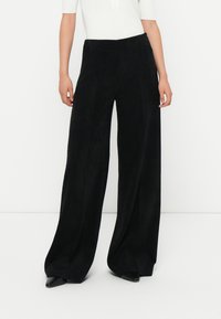 Schwarze weite Cordhosen mit strukturierter Oberfläche und hohem Bund. Die Kombination mit spitzen schwarzen Schuhen vollendet den Look.