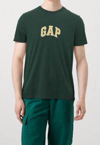 Hombre con camiseta oscura verde de GAP y pantalones cortos de carga en color aguamarina, de pie contra un fondo claro liso.