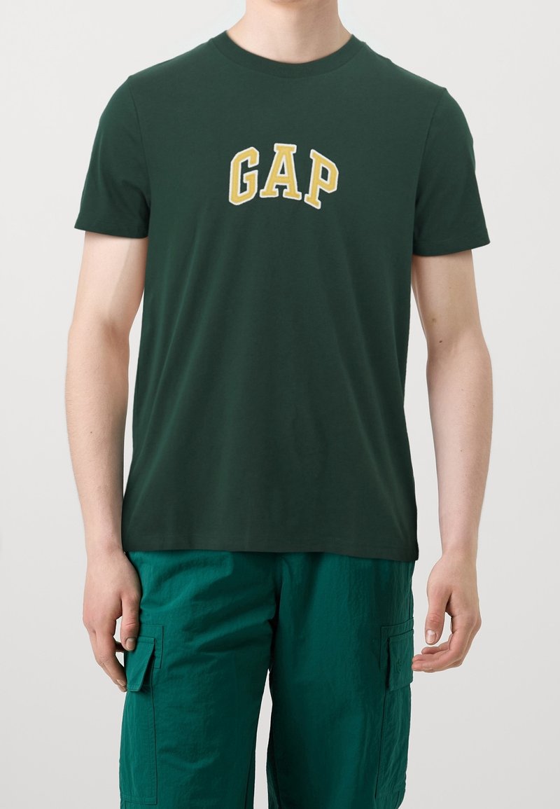 Hombre con camiseta oscura verde de GAP y pantalones cortos de carga en color aguamarina, de pie contra un fondo claro liso.