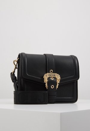 Bolso bandolera negro de cuero Versace Jeans Couture con hebilla ornamental dorada y correa de tela ajustable, apoyado sobre una superficie blanca.