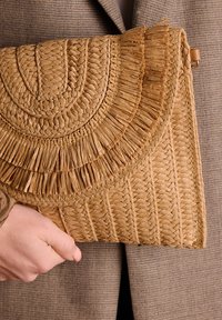 Geweven stro clutch met een gestructureerde oppervlakte, met een ronde, franjesdesign in natuurlijke beige tinten, vastgehouden door een hand tegen een bruine jas.