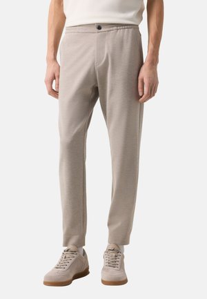 Mężczyzna ubrany w beżowe, dopasowane spodnie i beżowe sneakersy, stojący z rękami luźno opuszczonymi wzdłuż ciała.