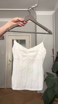 O mână ținând un umeraș din lemn cu un top fără mâneci din dantelă albă atârnând în interior, aproape de o ușă și de o plantă verde.