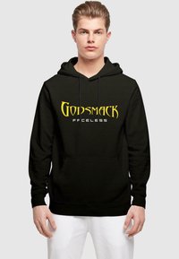 Crna hoodie sa žutim "GODSMACK" logom i natpisom "FFCELESS" ispod. Dizajnirana s prednjim džepom i kapuljačom s trakama. Model nosi bijele hlače.