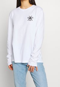 Frau trägt ein weißes Langarmshirt mit einem schwarzen asiatischen Symbol und einem grafischen Motiv von gekreuzten Schwertern auf der linken Brust, kombiniert mit hellblauen Jeans.