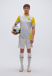 Φανέλα της Borussia Dortmund σε γκρι και κίτρινο χρώμα με γεωμετρικό σχέδιο, συνδυασμένη με λευκό σορτς, κάλτσες και πολύχρωμα παπούτσια ποδοσφαίρου, κρατώντας μια μπάλα.