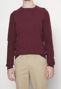 Burgunderfarbener Strickpullover mit Rundhalsausschnitt, gerippten Bündchen und Saum, aus strukturiertem Stoff gefertigt. Kombiniert mit hellbeigen Hosen.