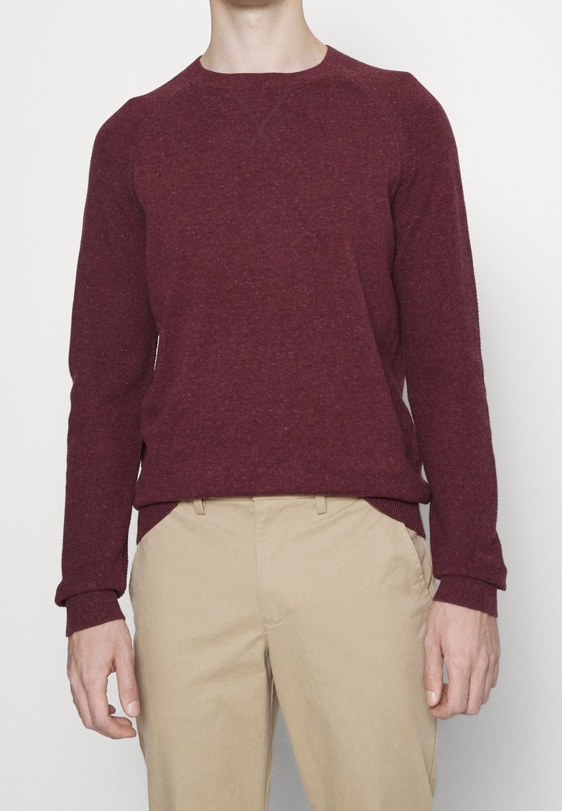 Burgunderfarbener Strickpullover mit Rundhalsausschnitt, gerippten Bündchen und Saum, aus strukturiertem Stoff gefertigt. Kombiniert mit hellbeigen Hosen.