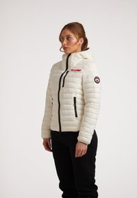 Giacca imbottita bianca con chiusura a zip, dettagli neri, cappuccio e logo sul petto e sul manica. Presenta cuciture a baffle orizzontali.