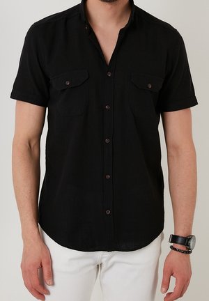 Zwart kortekraag overhemd met twee borstzakken, bruin knopen en een zachte textuur. Draagt een combinatie met witte broek en een horloge.
