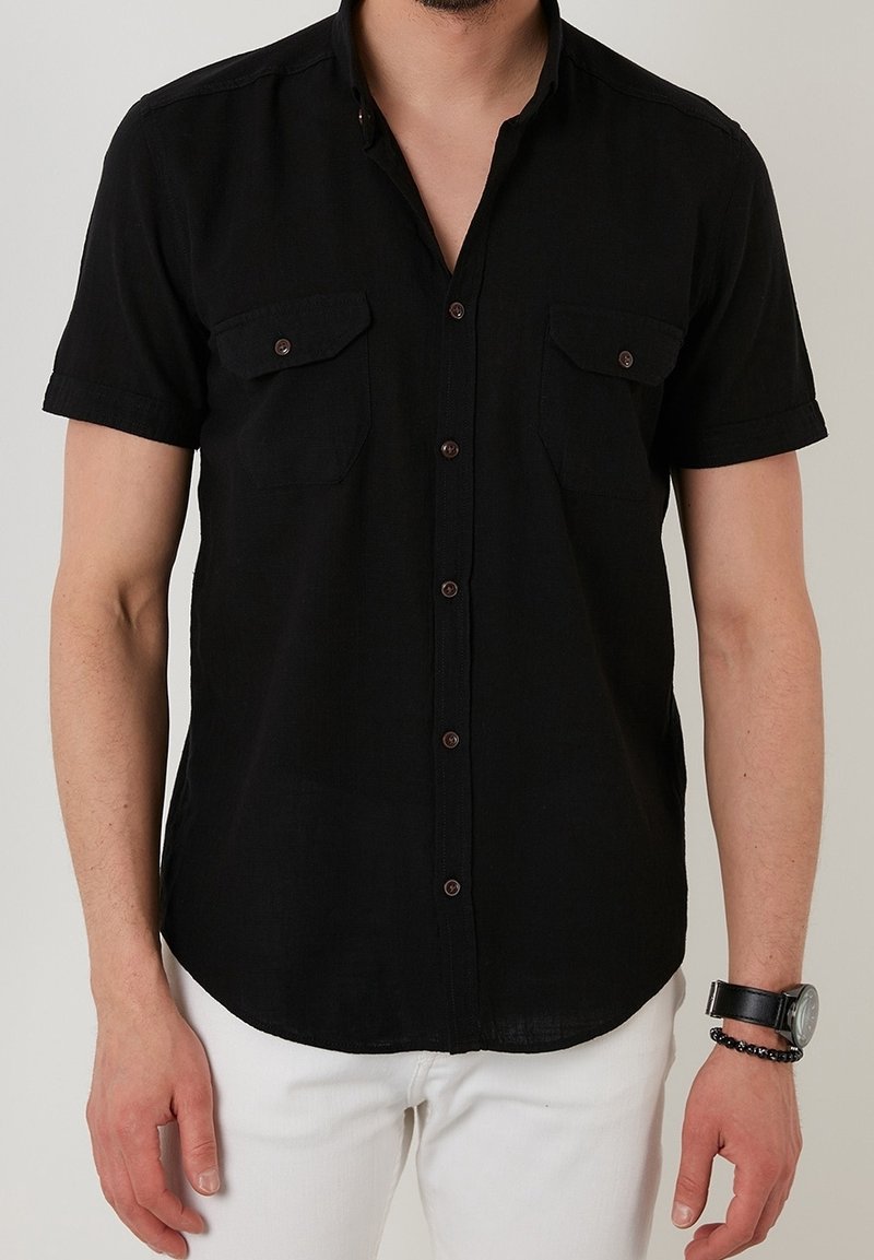 Kortärmad svart skjorta med knappar, två bröstfickor, bruna knappar och en mjuk textur. Bärs med vita byxor och en klocka.