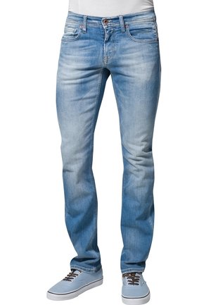 Jeans slim bleu clair portés avec des baskets bleu clair et une chemise blanche, montrés de la taille aux pieds sur un fond blanc.