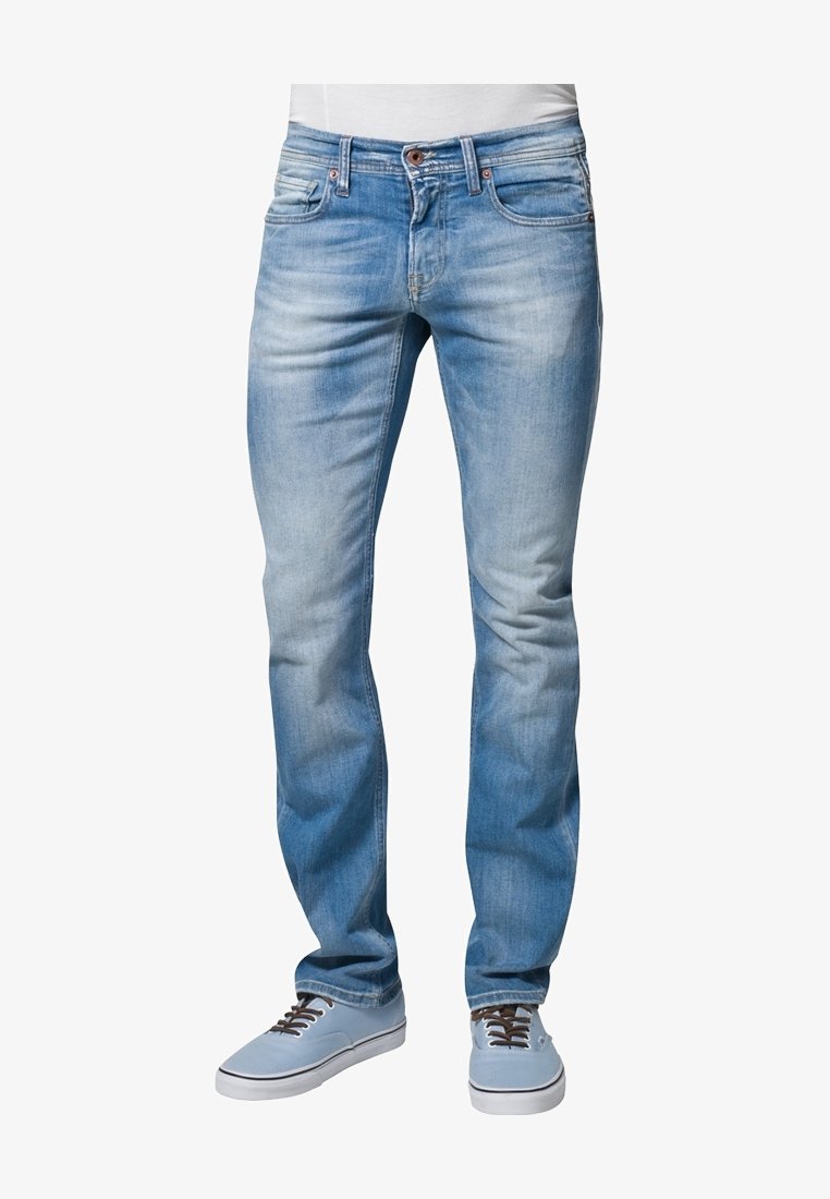 Jeans slim bleu clair portés avec des baskets bleu clair et une chemise blanche, montrés de la taille aux pieds sur un fond blanc.