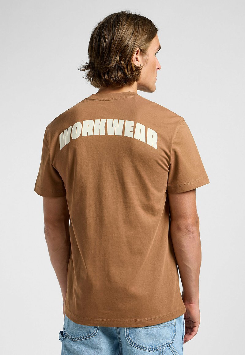 Brun bomull t-shirt med stora vita "WORKWEAR"-bokstäver på baksidan. Klassisk passform med korta ärmar och rundad halsringning.