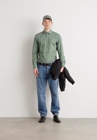 Cămașă verde cu nasturi, blugi albaștri, jachetă neagră în mână, pantofi negri, șapcă gri. Material neted, croială lejeră, design simplu. Fundal neutru.