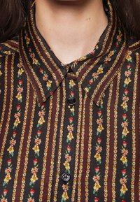 Gros plan d'une chemise marron boutonnée avec des rayures verticales et de petits motifs floraux jaunes, rouges et orange.